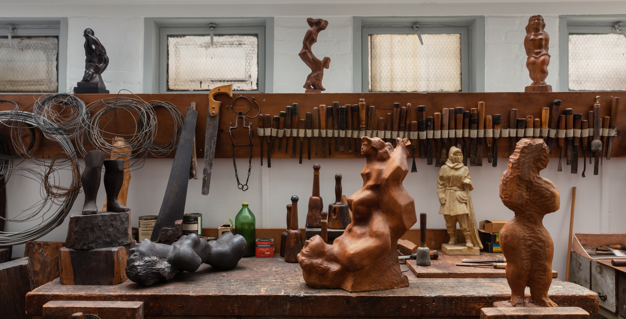 Photo of Chaim Gross' workbench. In the background there is a row of sculpting tools, wires, and small sculptures. In the foreground, on the workbench, there are a few larger wooden sculptures of the human figure.