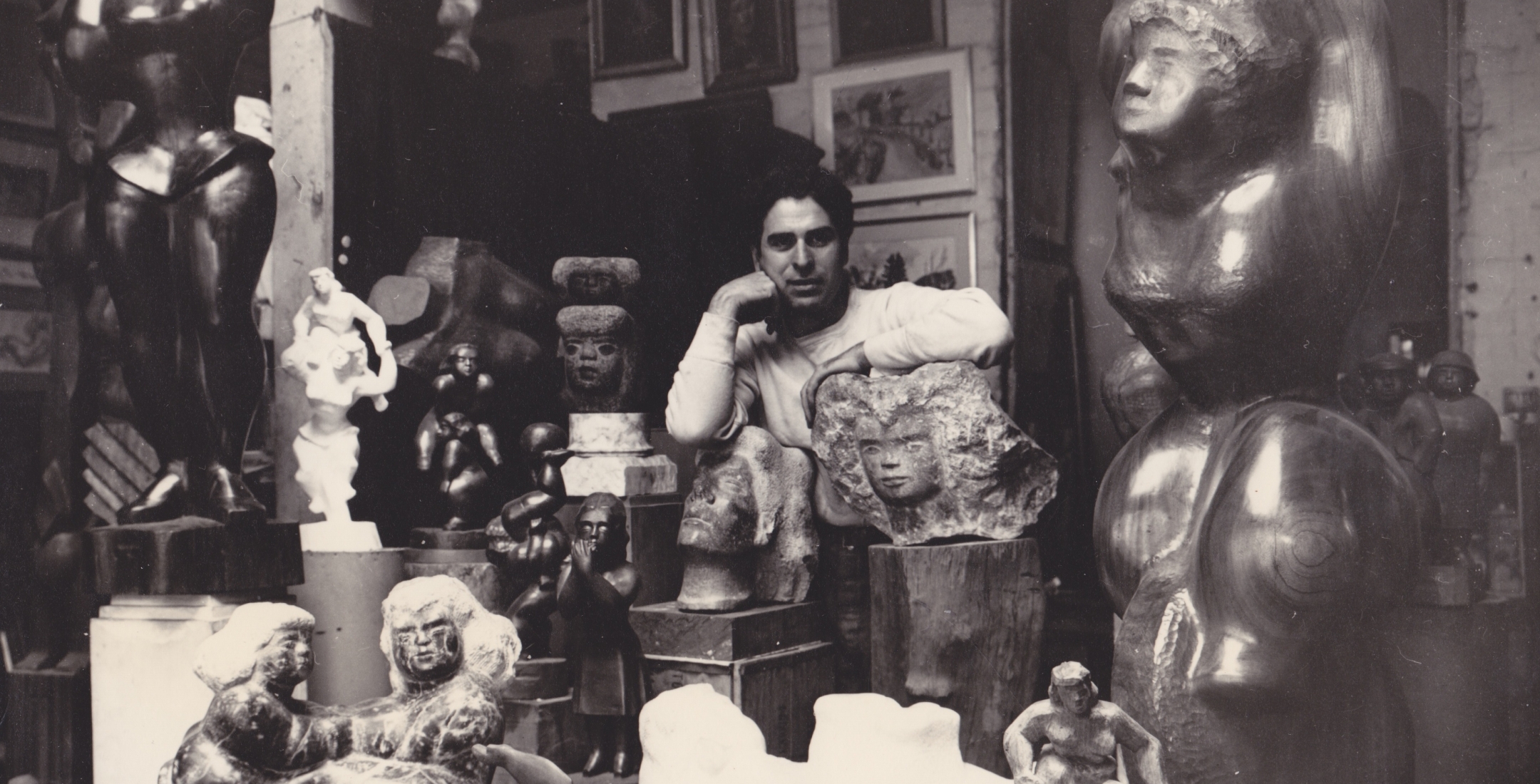 Black and white photo of a young Chaim Gross looking at the camera while surrounded by sculptures of various sizes and mediums.