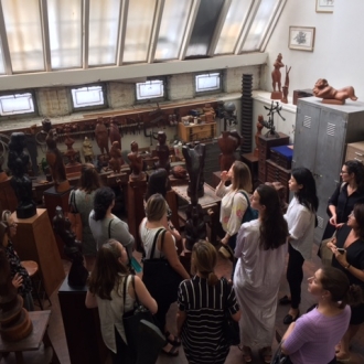 A large group of people crowds into the Chaim Gross Studio. They are surrounded by sculptures in various shades of wood and a large white skylight is above them. 