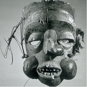 Mask, Ibibio. Mask made of wood, stain, reed, and fabric, replicating a human profile, with eyes carved out beneath the eyelids. The mouth is open, bearing teeth, and its bulbous cheeks and twisted nose greatly protrude from the mask. 