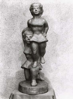 Black and white photo of a wooden sculpture. The sculpture depicts two female figures with one figure being lifted on the shoulder of the other. 