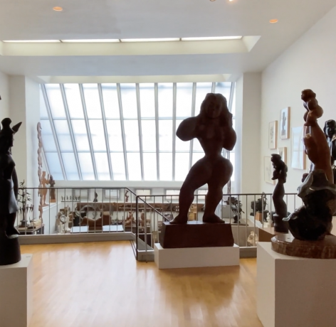 Photo of the top level of the 526 Laguardia Place double height studio space. The viewer stands on the top level, surrounded by sculptures positioned on white bases. One can see the skylight in the background, prints on the walls, as well as Chaim Gross' studio and workbench, complete with other works of his creation, below.