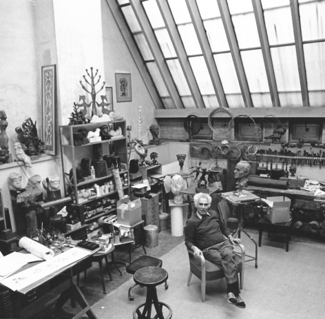 Black and white photo of Chaim Gross sitting in his studio surrounded by his art, his workbench, and his tools. The studio's large skylight looms above, at the top right corner of the photo.