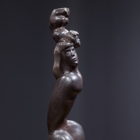 Photo of a wooden sculpture, I LOVE MY BABY, 1948, depicting a woman with long hair, crouched so that her legs are drawn close to her tall torso. She is supporting the figure of a baby, whose head touches hers but whose body is raised above its head. To the right of the sculpture are two framed ink sculpture studies for the same sculpture. The walls are faded navy.