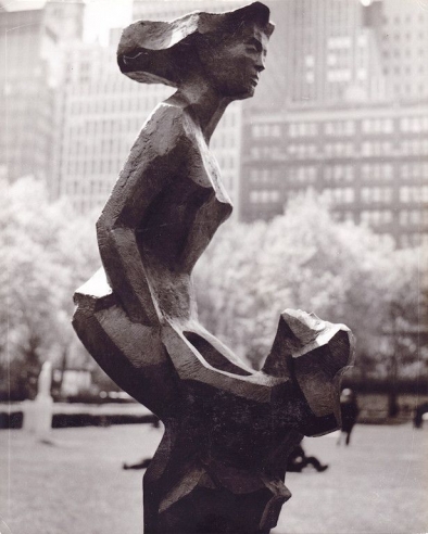 Geometric, wood sculpture of a tall woman, her hair in a stiff bulbous shape behind her, holding hands with a small girl gazing up at her. The legs of the woman are interconnected with the child, forming the base of the sculpture. 