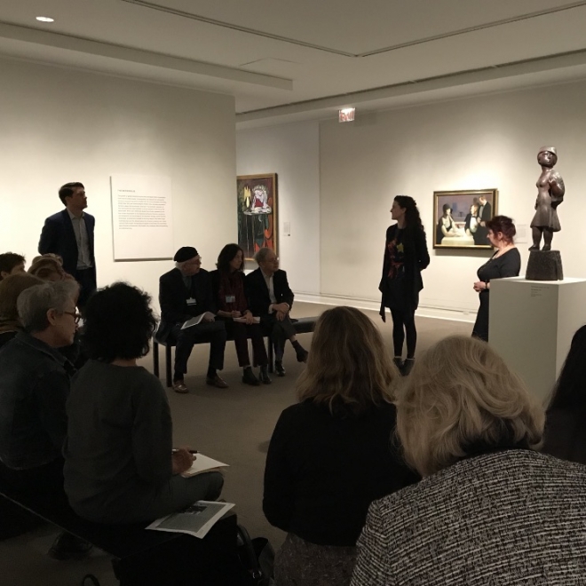 A group of adults sit in a white art gallery with low ceilings. Sasha Davis stands directly across from us in front of the people seated next to Mimi Gross. To the left of them is a small sculpture of a female figure with condensed proportions. In background there are two large framed paintings in a hallway facing left sideways out of the frame of the photo. 