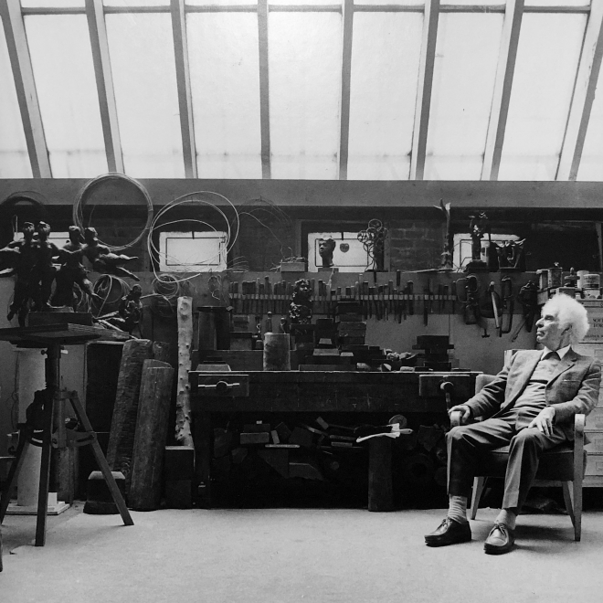 A black and white photo of an elderly Chaim Gross with prominent fluffy white hair wearing a suit sits to the left of us. He is facing sideways away from us, looking at his large collection of tools, work bench, and a sculpture of a cluster of female forms in playful motion. 