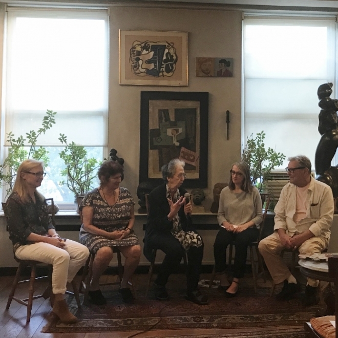 Three older adults and two adults are seated in a half moon shape, all facing an older person who is speaking into a microphone at the center. Behind them are two large windows with several pieces of framed artwork between them, as well as a few sculptures and plants. 