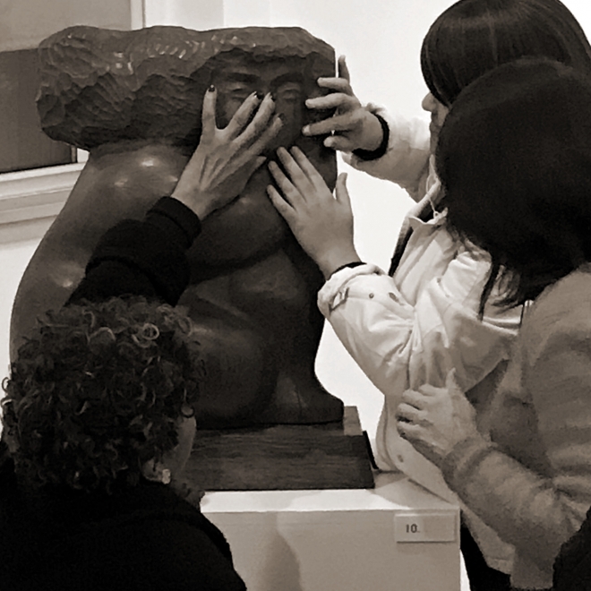 Close up black and white photo of three adults with dark hair touching the face of a large, wide, wood sculpture of a humanoid subject with a blocky, smooth body shape. 