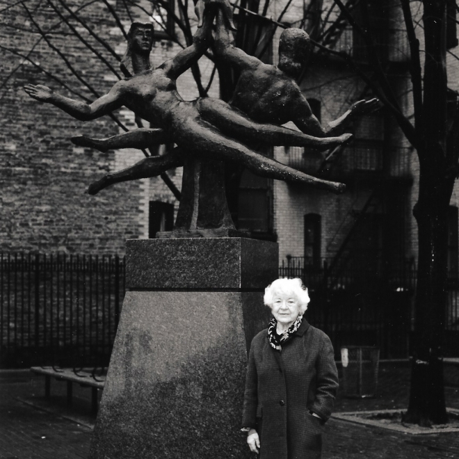 A black and white photo of an elderly Renee Gross in a dark winter coat with bright white hair standing in front of a large outdoor sculpture. The sculpture features two identical female forms laying sideways with their left hands raised and touching. In the background of the photo you can see apartment buildings made of brick. 
