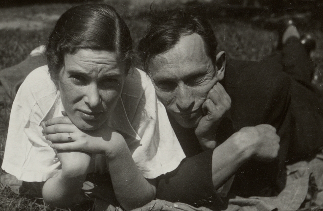 Black and white photograph of a man and woman lying on their stomachs in the grass. They are both resting their chins on their hands, looking directly at the camera.