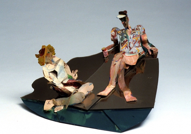 A sculpture of an adult man and a woman made of thin, brightly painted material sits on a dark organic shaped base. The woman has blonde hair and is turned upwards to face the male sculpture who is sitting up higher in the composition. The man looks downwards, his face in shadow. 